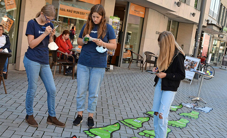 Drei Schülerinnen stehen in einer Fußgängerzone und nutzen die großen Fußaufkleber am Boden, um den ökologische Fußabdruck zu ermitteln.