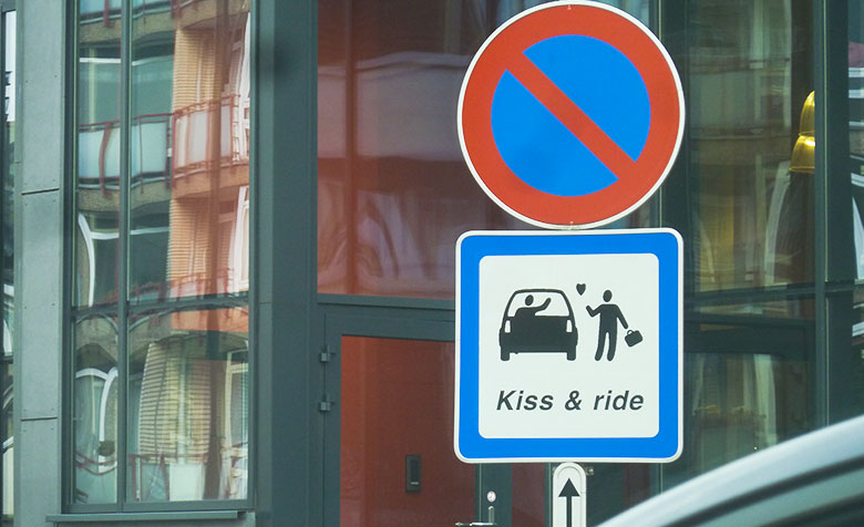 Unter ein Parkverbot-Schild in Brüssel wurde ein weiteres Schild angebracht, auf dem steht: „Kiss & Ride“ und ein Figürchen einem zweiten im Auto sitzenden zuwinkt. Zwischen den beiden ist ein Herz.