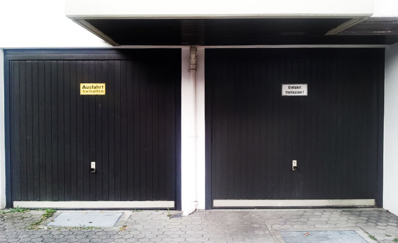 Zwei nebeneinanderstehende Garagentore sind beschriftet. Die eine mit „Ausfahrt freihalten“, die daneben mit „Einfahrt freilassen!“