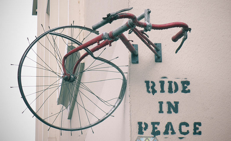 An einer Hauswand in Paris ist ein zerbeultes Vorderrad eines Fahrrads angeschraubt. Darunter wurde „Ride in Peace“ an die Wand gesprüht.