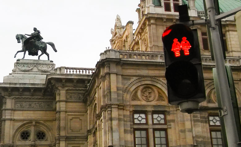 Das grüne Licht einer Ampel zeigt zwei händchenhaltende Frauen mit einem Herz zwischen den beiden Köpfen. Im Hintergrund ist die Wiener Staatsoper.