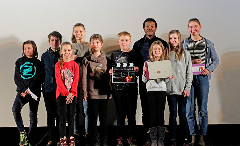 Die Kinder, die den Film „Glück im Unglück“ gedreht haben, stehen mit einer Filmklappe und Urkunde auf einer Bühne.