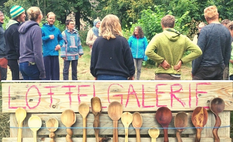 Viele Holzlöffel hängen an einem Holzbrett auf welchem "Löffelgalerie" steht. Dahinter stehen Menschen in einem Kreis.